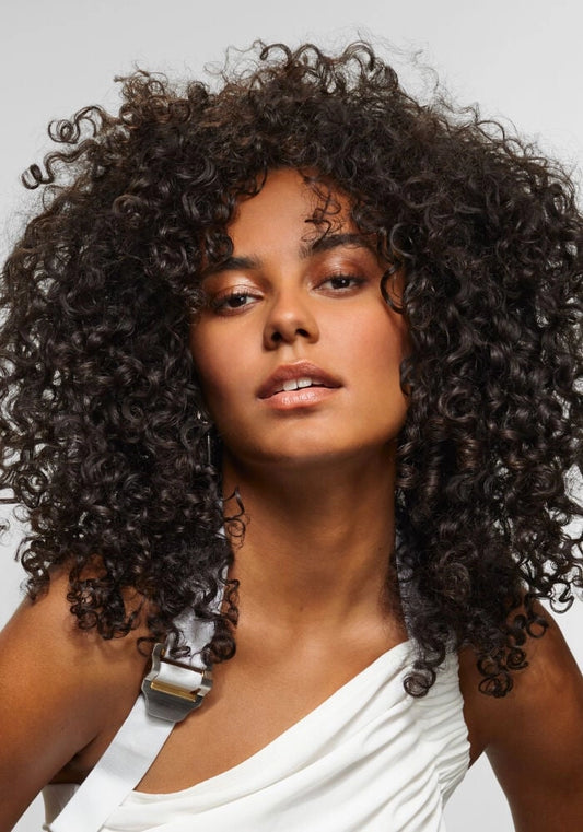 A close-up portrait of a woman with voluminous, tightly curled dark brown hair, wearing a white draped top against a light gray background.