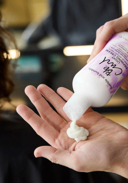 A close-up of a person pouring white hair-denying cream from a purple and white bottle into their palm, with the brand name 'The Curl' visible on the label.