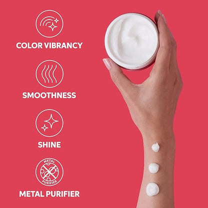 A hand holds an open white cosmetic jar against a bright pink background, with three dollops of white cream on the wrist. White text on the left lists benefits: color vibrancy, smoothness, shine, and metal purifier.